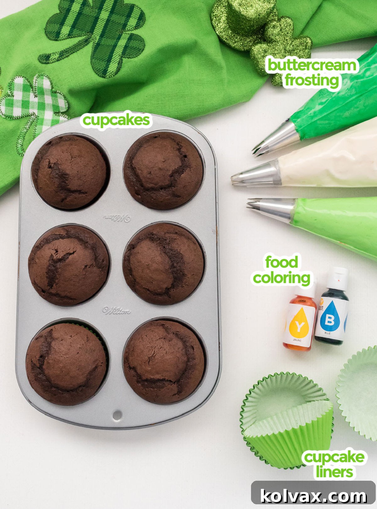 A collection of ingredients needed to make St. Patrick's Day Cupcakes, including baked chocolate cupcakes, a bowl of homemade buttercream frosting, green and blue food coloring, and green cupcake liners.