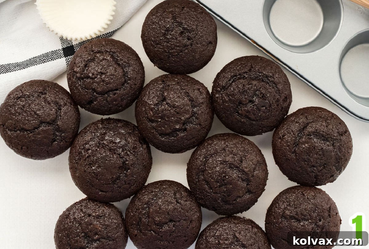 An overhead shot of 12 freshly baked chocolate cupcakes in green liners, cooling on a white table next to an empty cupcake pan, ready for frosting.