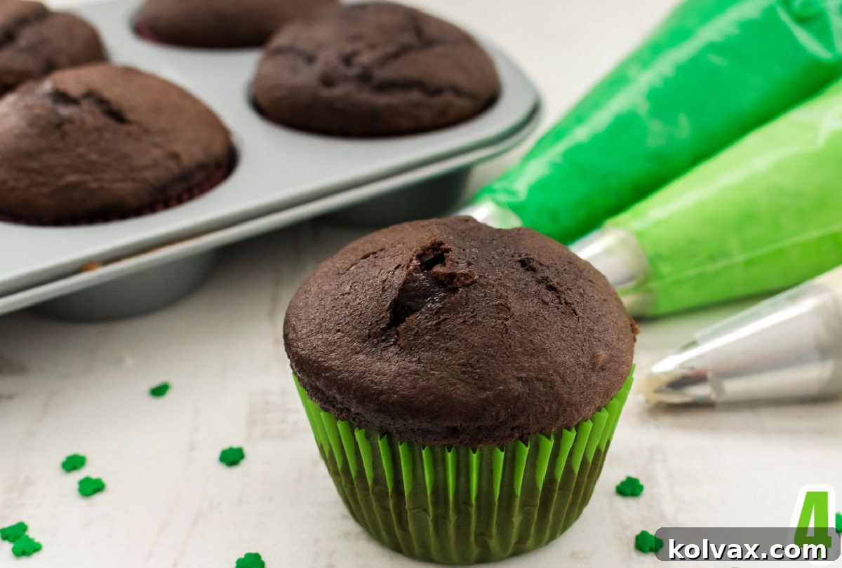 A single chocolate cupcake in a green liner sits in the foreground, with a cupcake tin full of other cupcakes and three piping bags filled with different colored frostings (dark green, light green, white) visible in the background.