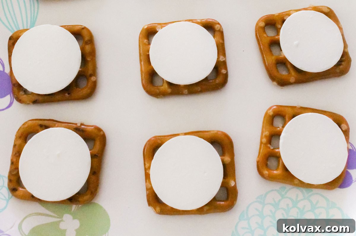 A microwave-safe paper plate holds six pretzels, each topped with a pristine white chocolate melting wafer, ready for the microwave.