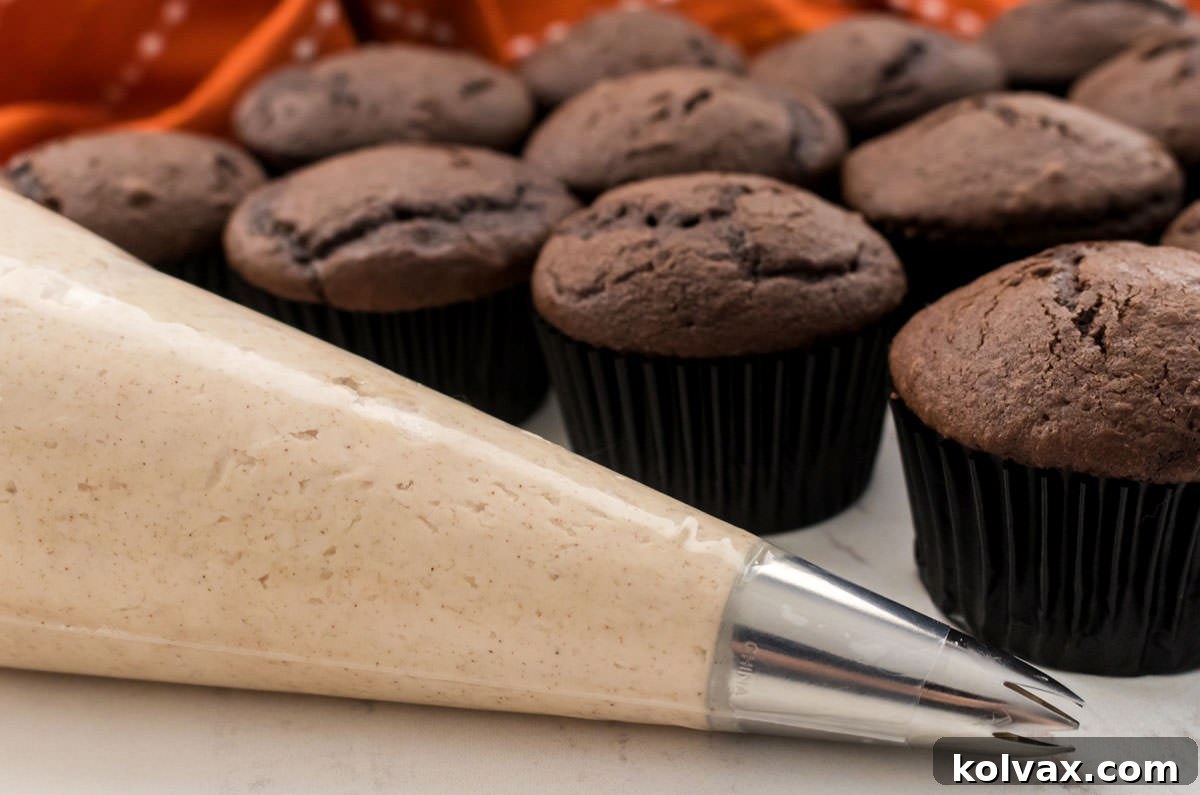 Heavenly Cinnamon Buttercream 6 A piping bag elegantly filled with smooth Cinnamon Frosting, positioned in front of a tray of unfrosted chocolate cupcakes. This image suggests the delicious potential of decorating baked goods with this homemade frosting.