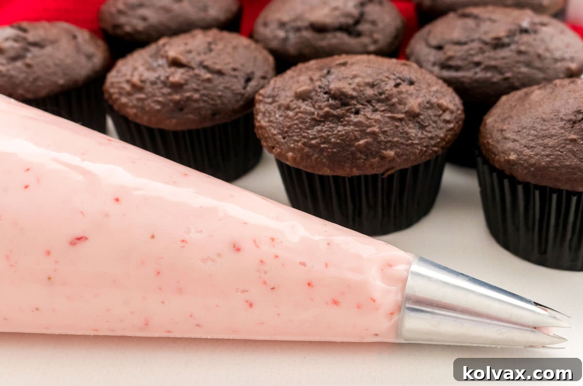 A decorating bag filled with pink homemade strawberry frosting sits on a white surface, ready to frost a batch of unfrosted chocolate cupcakes in the background.