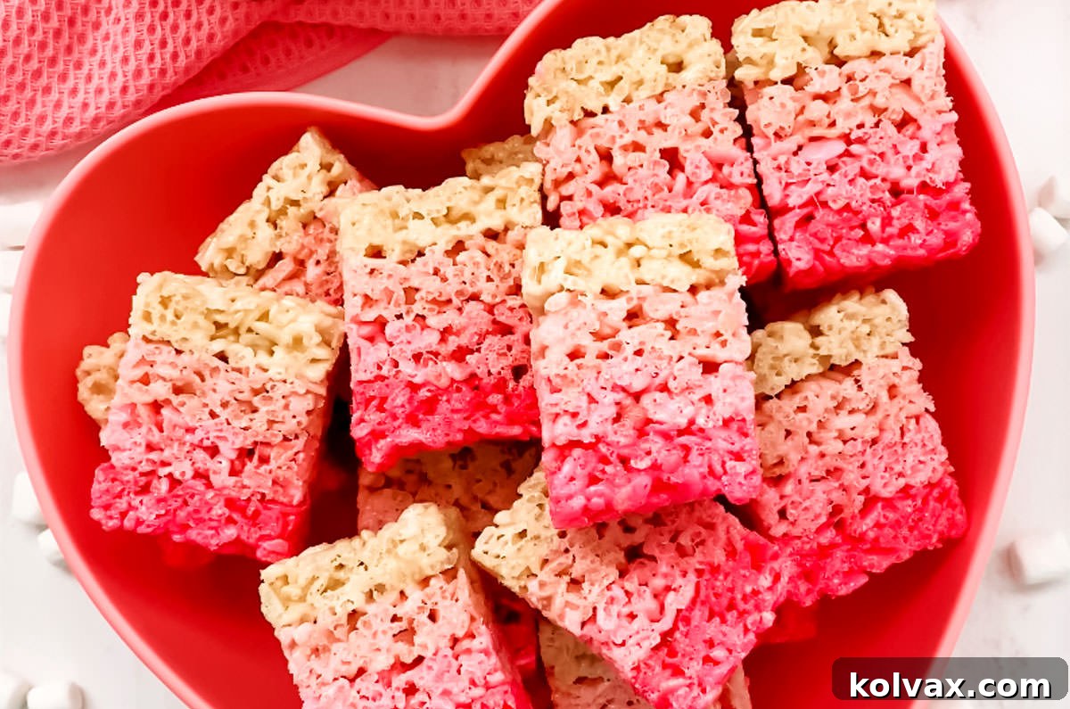 Close-up of a batch of perfectly layered Ombre Rice Krispie Treats, displaying a beautiful pink gradient, nestled in a heart-shaped pink bowl on a clean white table, ready to be enjoyed.