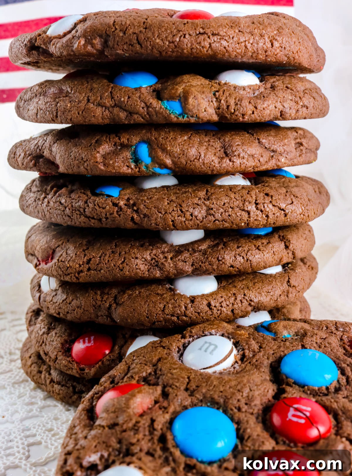 Closeup on a tall stack of Chocolate Red White and Blue M&M Cookies sitting on a white linen in front of a white background.
