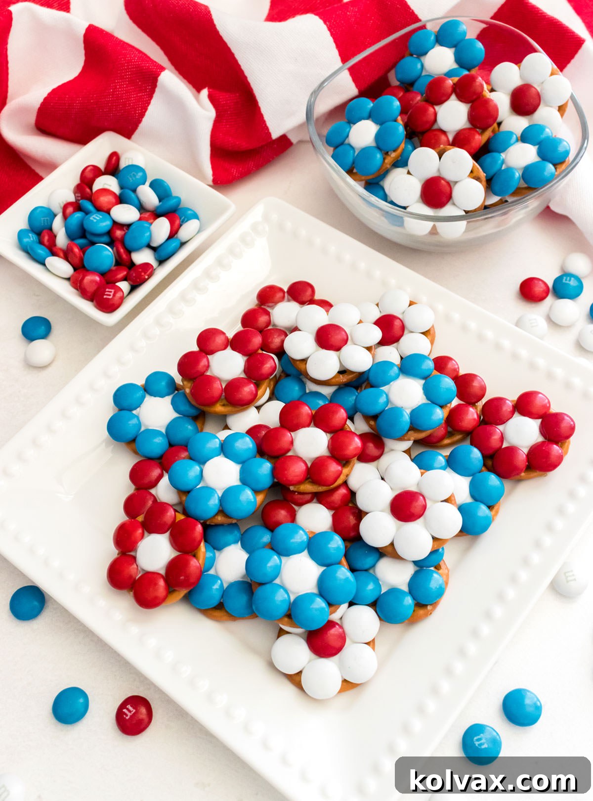 Closeup on a dessert plate filled with Patriotic Flower Pretzel Bites sitting on a white table with an American Flag Kitchen Towel and a ramekin filled with Red, White and Blue M&M's.