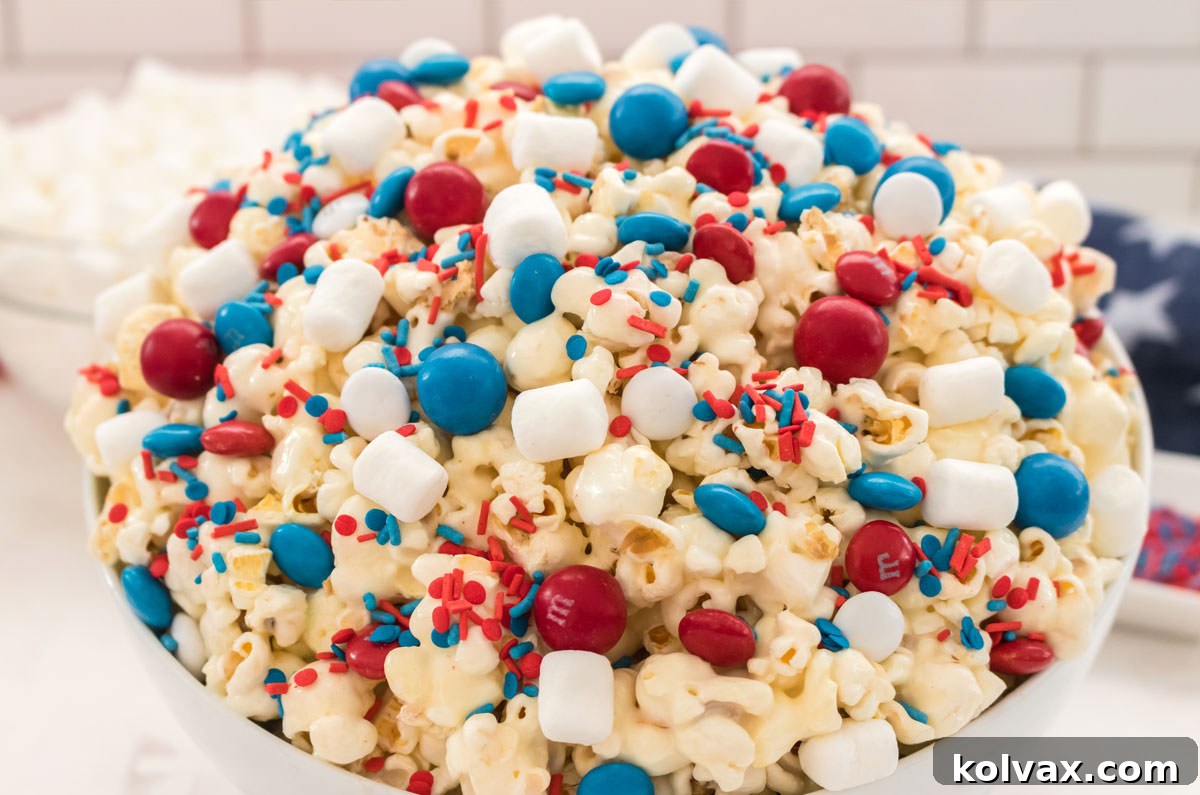 A close-up of a large white serving bowl brimming with colorful Patriotic Popcorn. The popcorn features a marshmallow coating, red, white, and blue candies, and sprinkles. It sits on a white table with a blurry background of marshmallows and an American flag, emphasizing its festive theme.