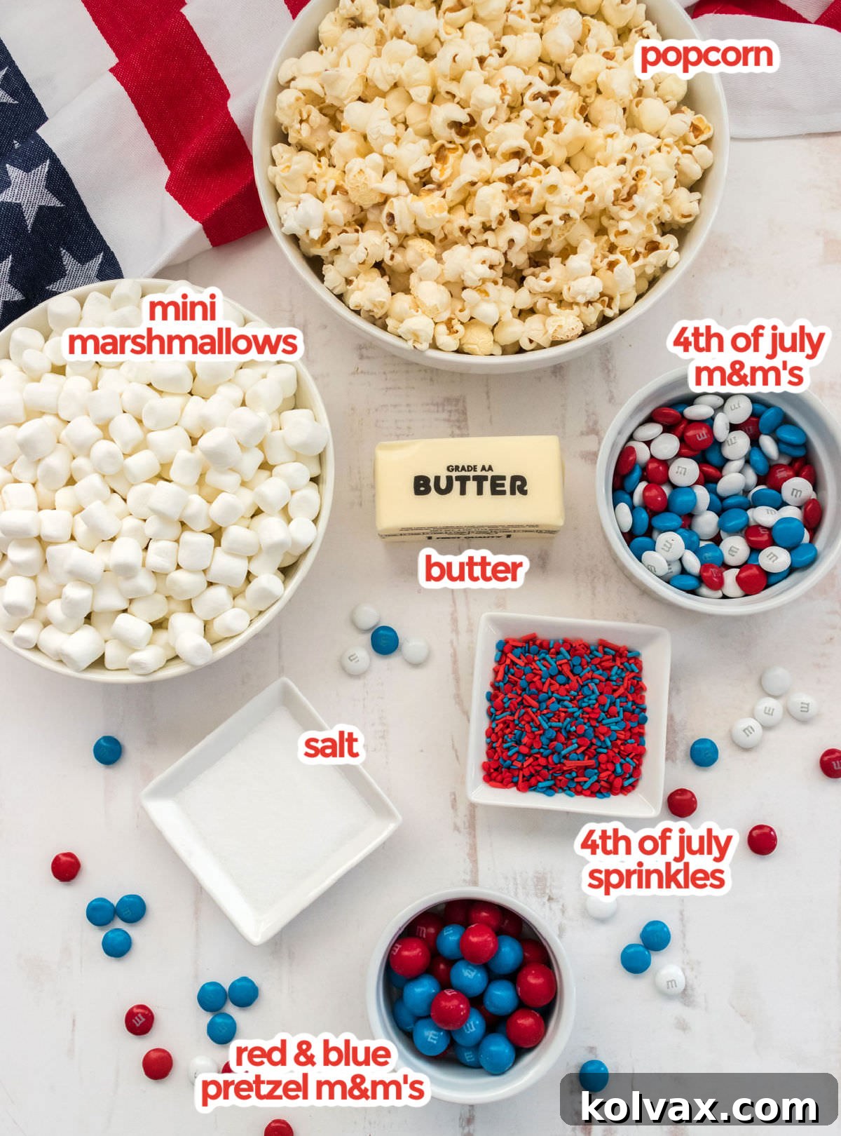 All the vibrant ingredients laid out for making Patriotic Popcorn, including freshly popped kernels, mini marshmallows, patriotic M&M's, rich butter, a pinch of salt, and colorful sprinkles. A bag of Pretzel M&M's is also visible, hinting at an extra crunchy and sweet-salty addition.