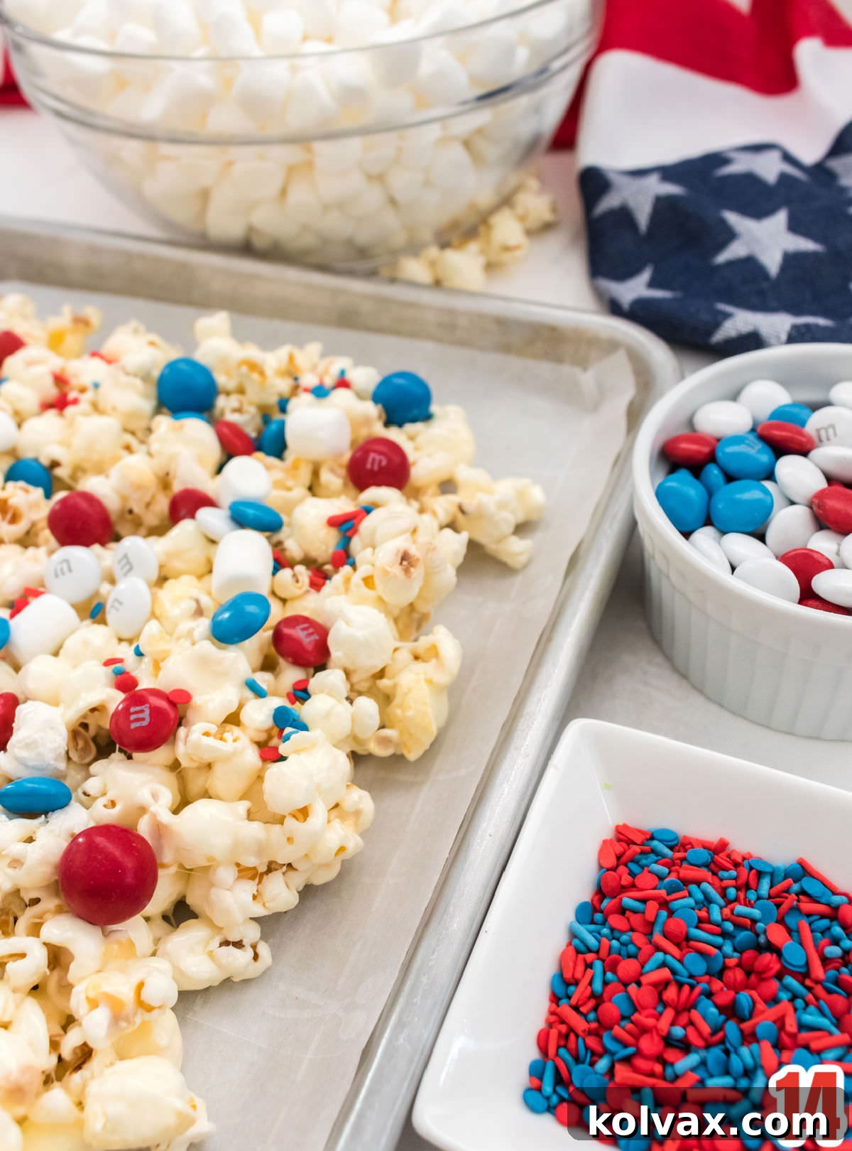Patriotic Popcorn spread on a cookie sheet, surrounded by bowls of colorful candy mix-ins: an array of sprinkles, mini marshmallows, and patriotic red, white, and blue M&M's. The scene showcases the final decorative touches that elevate this festive treat.