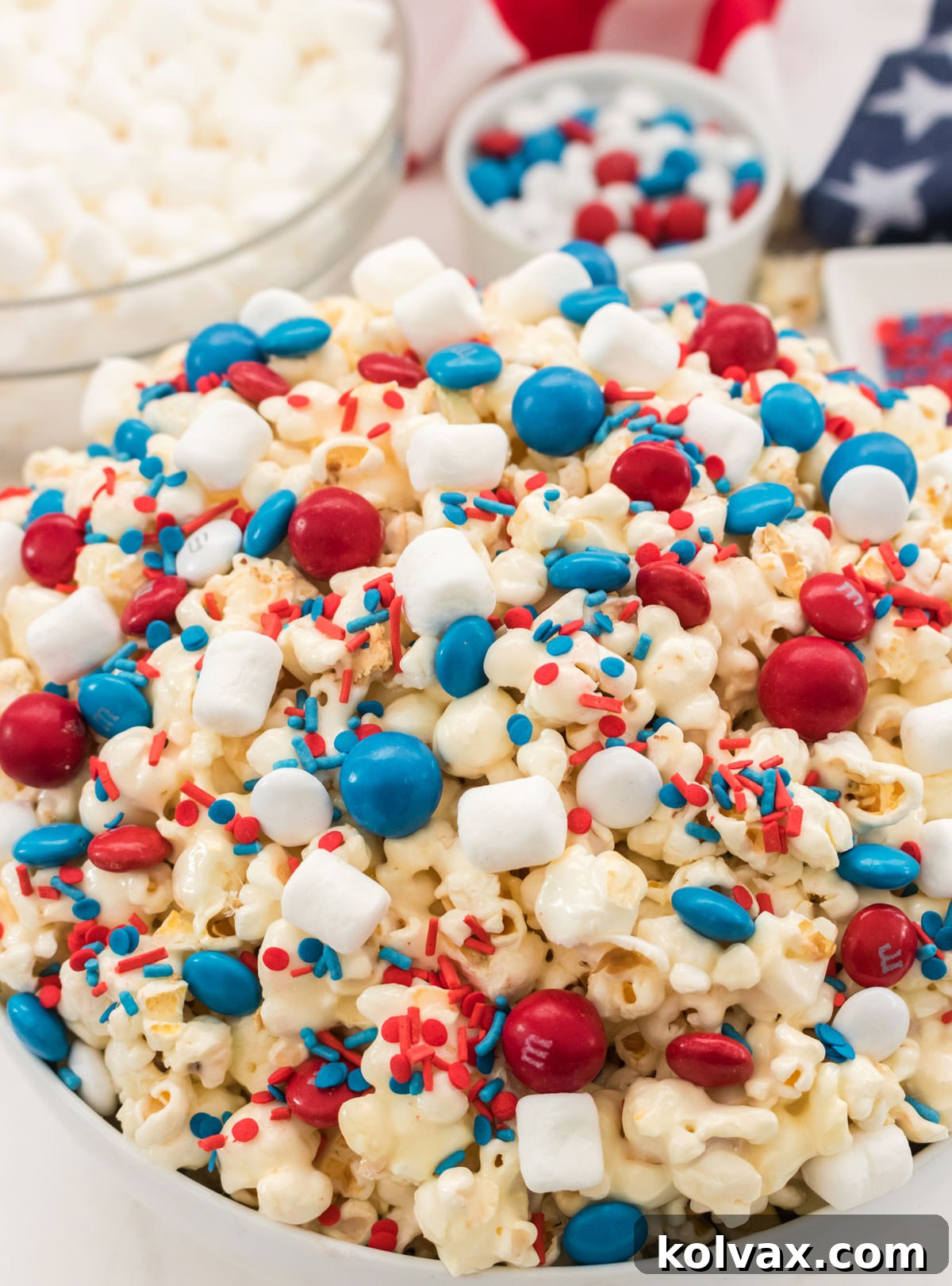 A vibrant close-up of a large white serving bowl overflowing with Patriotic Popcorn, featuring a delightful mix of marshmallow-coated kernels, red, white, and blue M&M's, and sprinkles. Behind it, a clear bowl of fluffy marshmallows and a ramekin of M&M's enhance the festive backdrop, perfect for any holiday celebration.