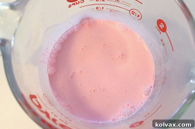 A small drop of pink food coloring being added to heavy whipping cream in a measuring cup, the first step for colorful homemade whipped cream.