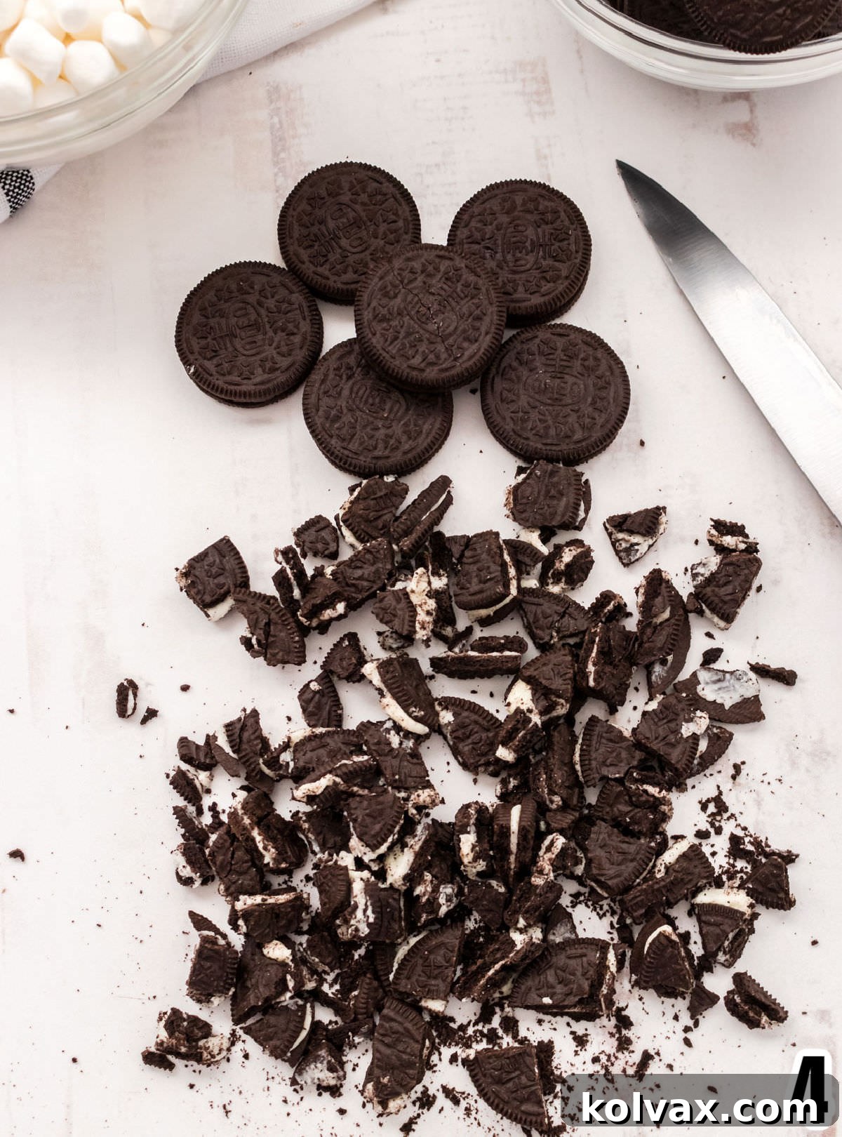 Midnight Cookie Pop 5 Close-up view of roughly chopped Oreo Cookies on a white cutting surface, with whole Oreos and a kitchen knife visible in the background, illustrating the preparation step.