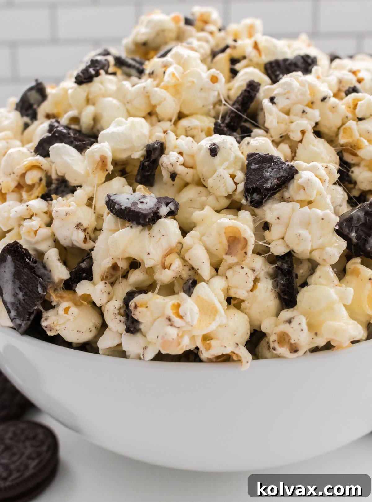 Midnight Cookie Pop 9 Closeup shot of a pristine white serving bowl overflowing with homemade Oreo Popcorn, artfully arranged on a white table and beautifully garnished with additional whole Oreo Cookies around the base, ready for serving.