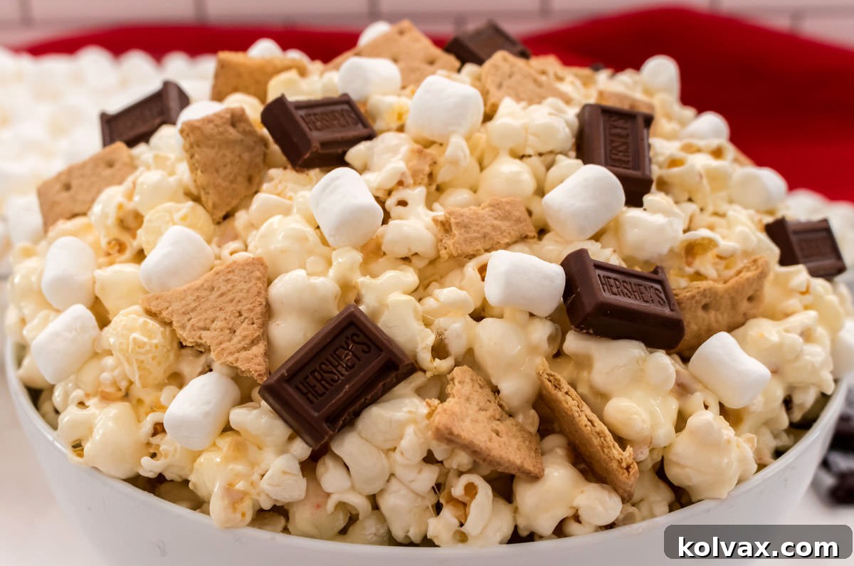 Campfire Crunch Popcorn 2 Closeup on a white bowl filled with S'mores Popcorn sitting in front of a bowl of mini marshmallows and a red kitchen towel, perfectly showcasing the sweet and salty treat.