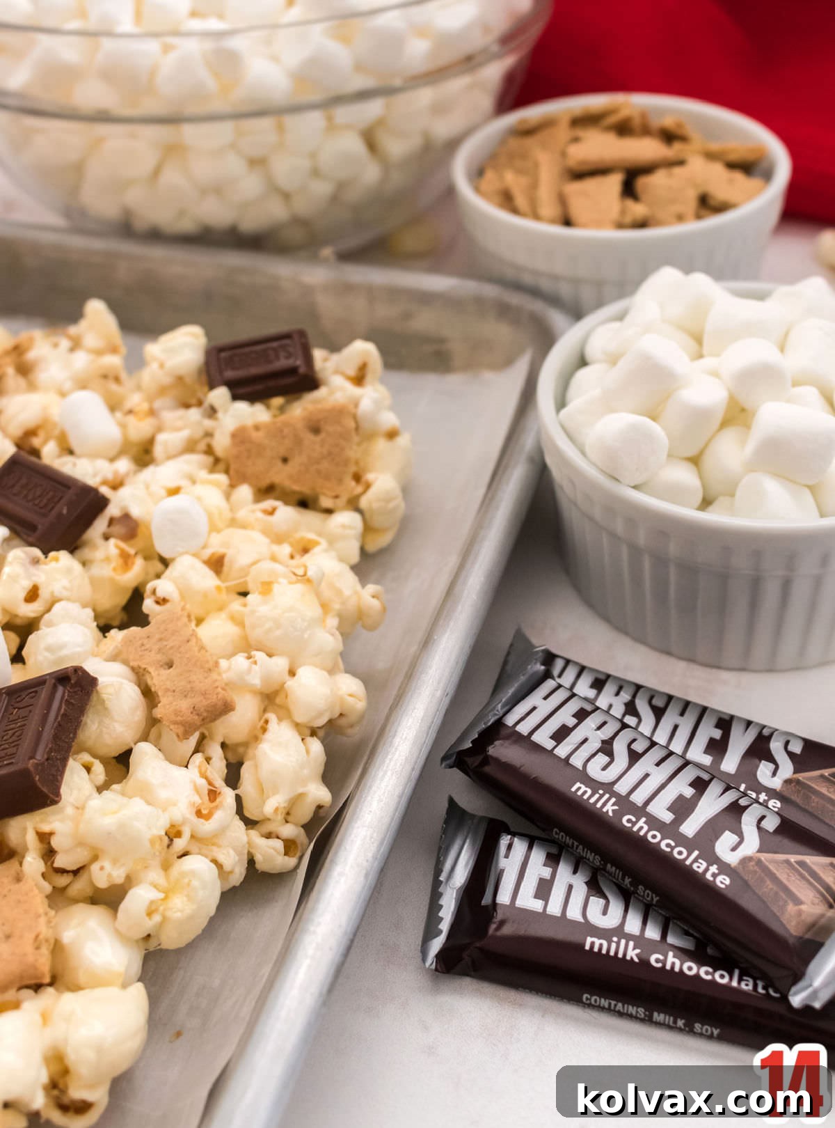 Campfire Crunch Popcorn 8 Close-up on a cookie sheet filled with S'mores Popcorn surrounded by ramekins filled with Mini Hershey's Milk Chocolate Bars, Mini Marshmallows, and Graham Cracker pieces, ready to be added.