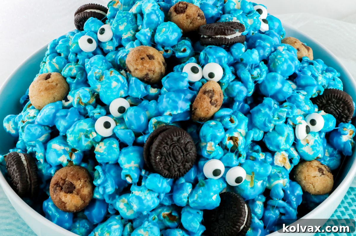 Closeup of a white serving bowl filled with vibrant blue Cookie Monster Popcorn, adorned with mini chocolate chip cookies and candy eyes, sitting on a blue placemat against a soft white background. This sweet and salty snack is perfect for parties or a fun movie night.