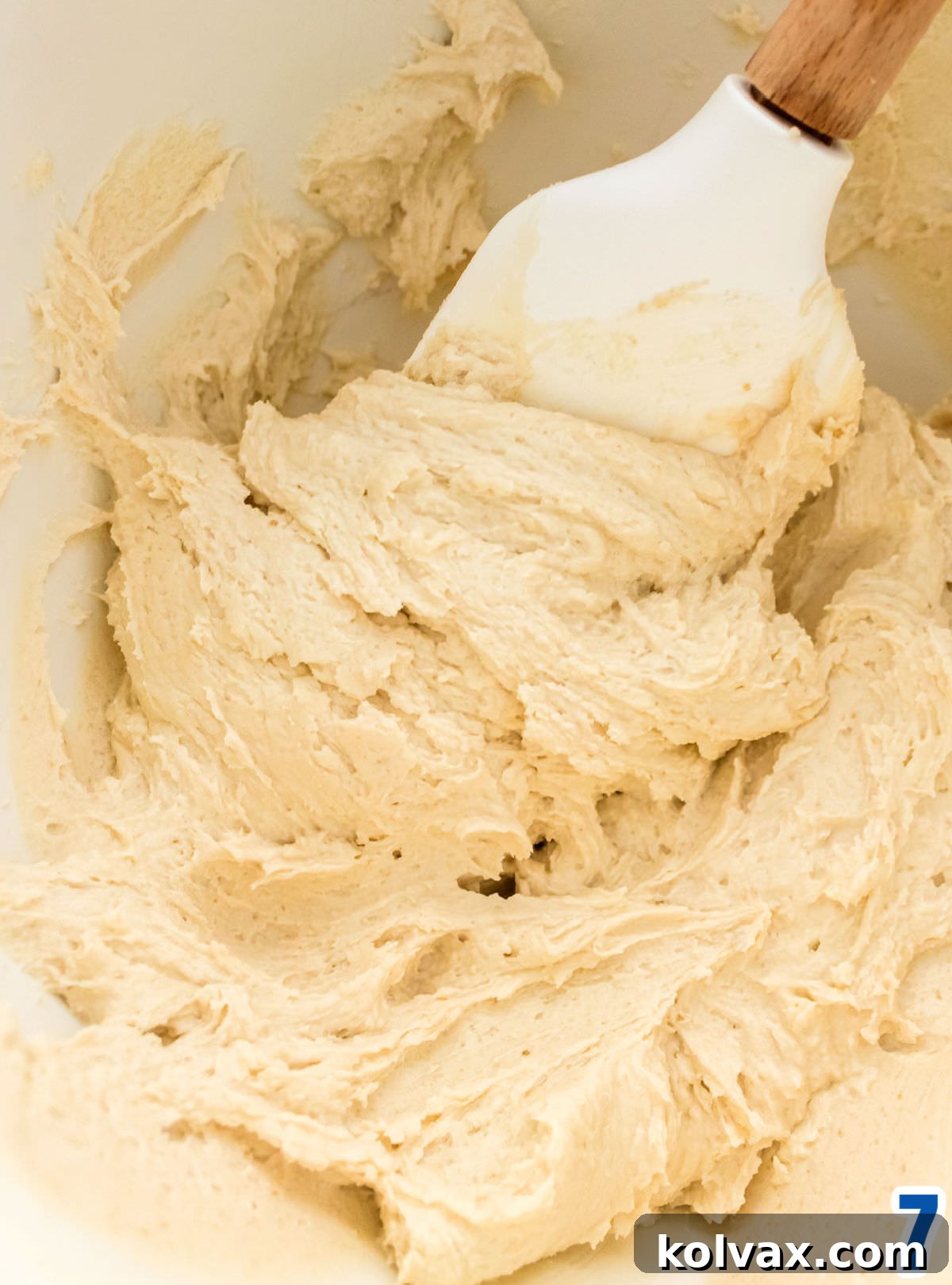 A close-up view of a pristine white mixing bowl filled with freshly made, smooth Brown Sugar Buttercream Frosting, ready for decorating, with a white wooden spatula resting nearby.