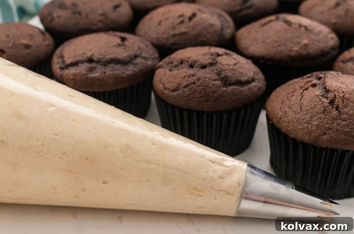 A decorating bag, neatly filled with creamy Brown Sugar Frosting, positioned in front of a tray of unfrosted chocolate cupcakes, awaiting their sweet adornment.