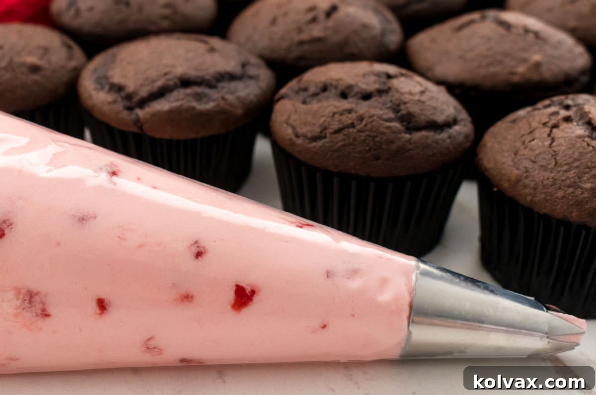 A piping bag filled with vibrant Maraschino Cherry Frosting, poised in front of a platter of unfrosted chocolate cupcakes, ready for decoration.