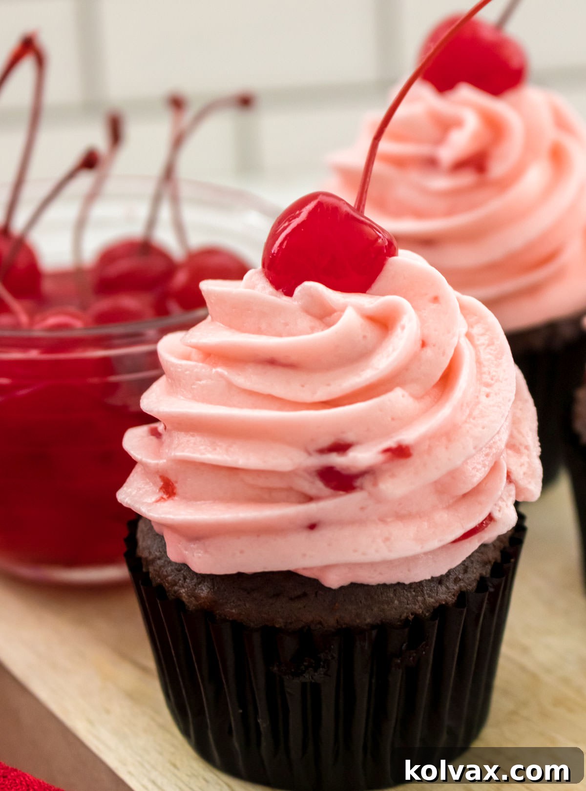 A close-up of a chocolate cupcake generously frosted with The Best Maraschino Cherry Buttercream Frosting, garnished with a whole maraschino cherry, next to a bowl of cherries.