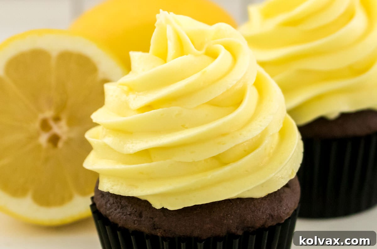 Two chocolate cupcakes frosted with the Best Lemon Buttercream Frosting, garnished with lemon zest, sitting next to a fresh lemon slice. The frosting is light yellow and perfectly piped.