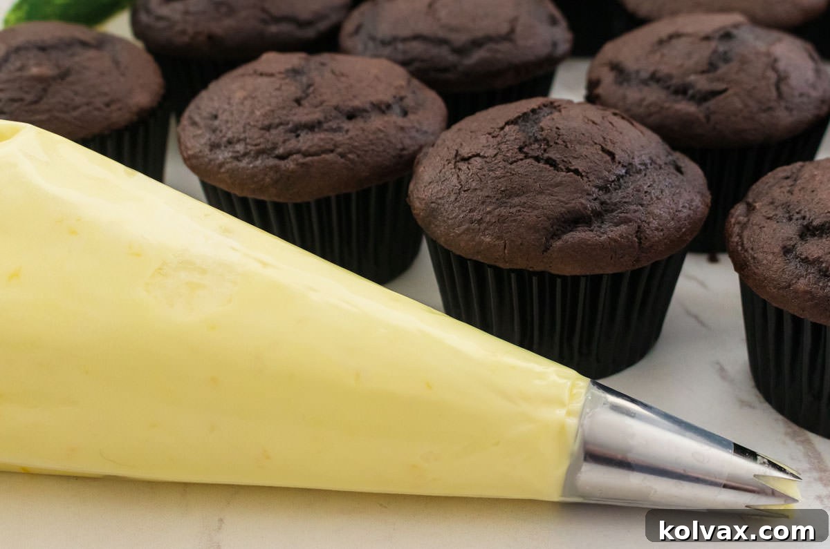 A decorating bag filled with light yellow Lemon Buttercream Frosting, ready to be piped, sitting in front of a tray of unfrosted chocolate cupcakes.