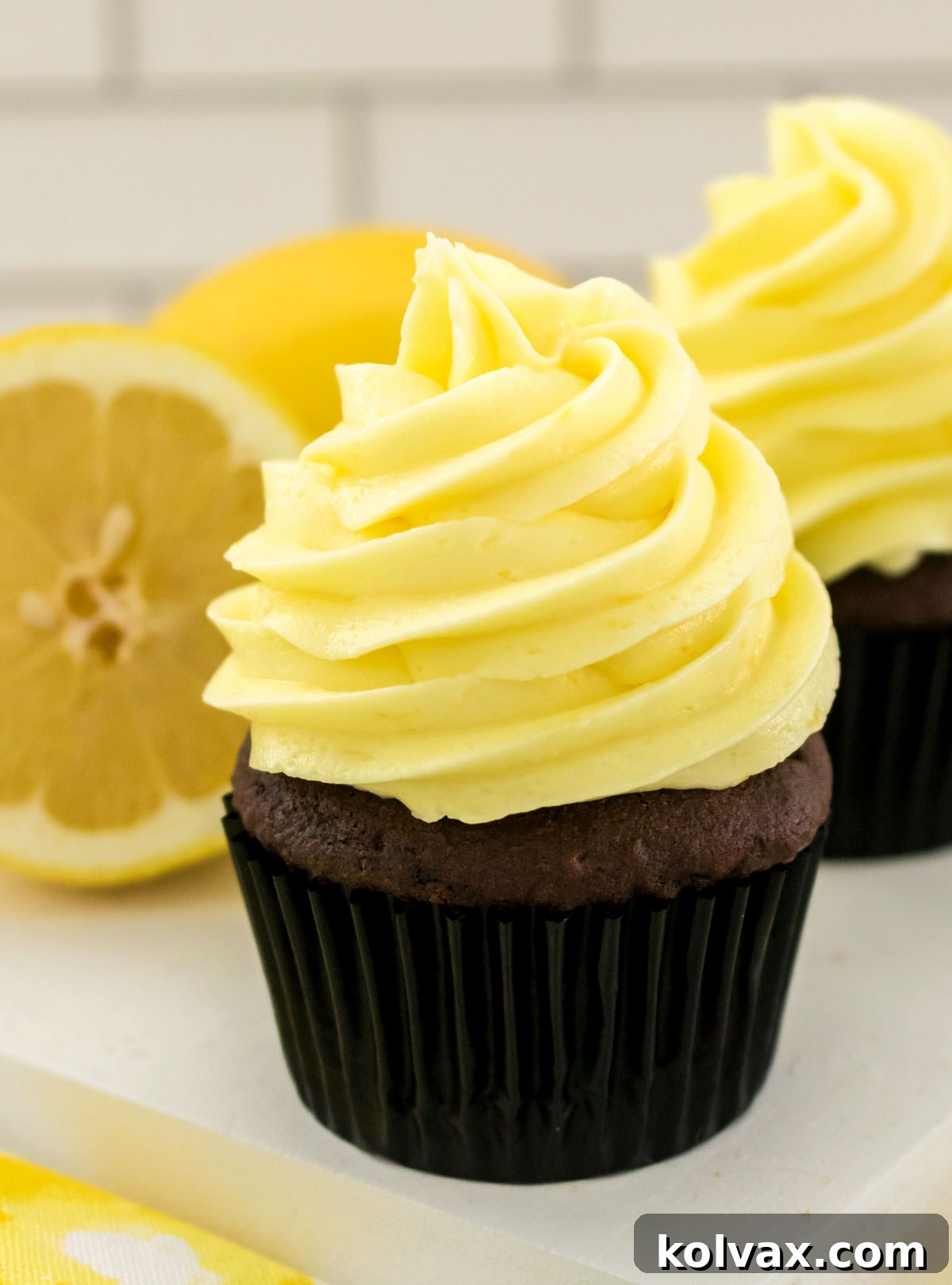 A perfectly frosted chocolate cupcake with The Best Lemon Buttercream Frosting, elegantly placed on a white platter next to a fresh lemon slice, ready to be enjoyed.