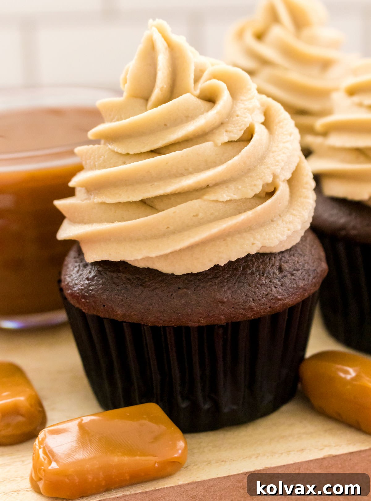 Closeup on two chocolate cupcakes generously frosted with The Best Caramel Buttercream Frosting, beautifully presented next to a bowl of caramel sauce.