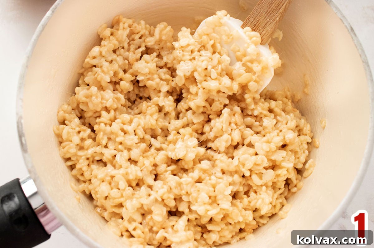 A close-up view of a pristine white pan containing a warm, gooey Rice Krispie Treat mixture, ready for the next step, with a white wooden spoon resting within it.