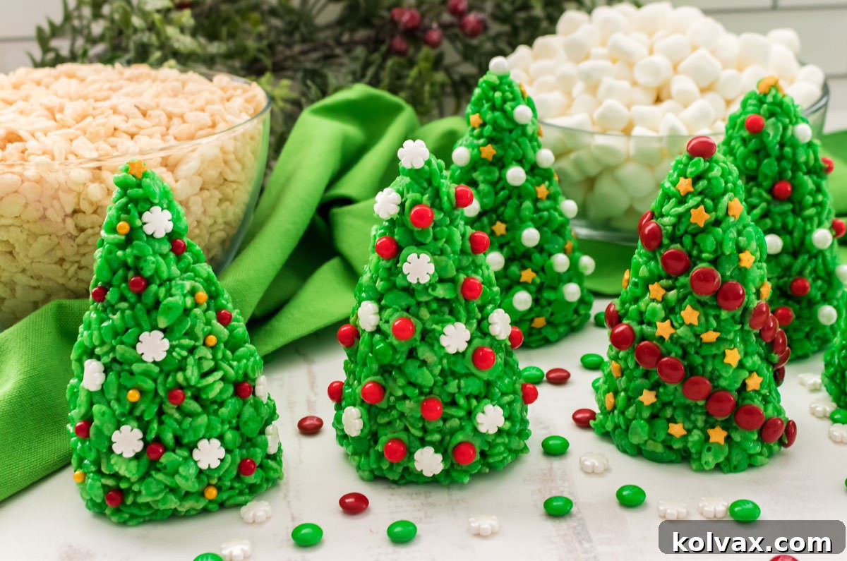 Closeup of five vibrant green Christmas Tree Rice Krispie Treats adorned with sprinkles and M&M's, sitting on a white table with bowls of mini marshmallows and rice krispies in the background.