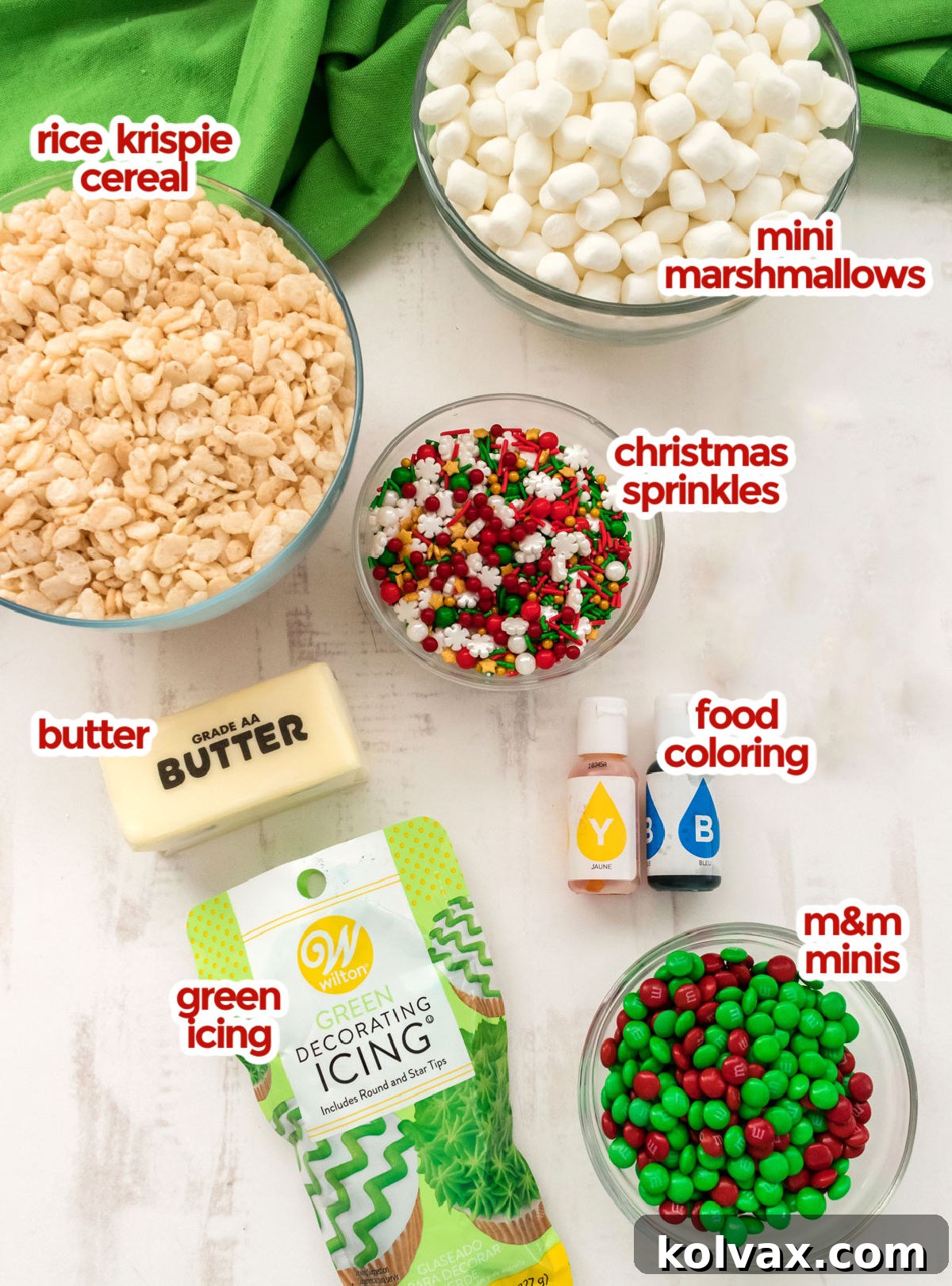 A curated display of all the necessary ingredients for Christmas Tree Rice Krispie Treats, including a bag of mini marshmallows, Rice Krispie cereal, a stick of butter, bottles of food coloring, a jar of festive sprinkles, and a tube of green icing.