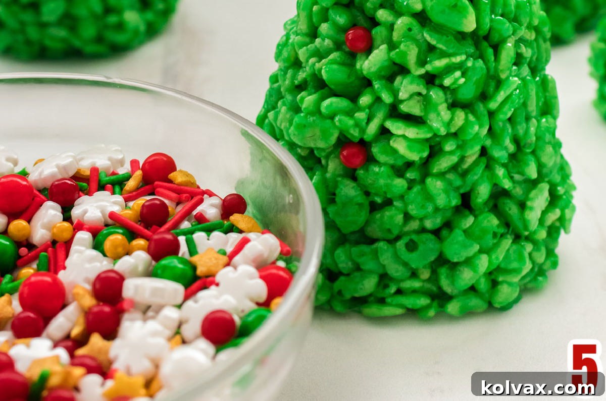 A beautifully decorated Christmas Tree Rice Krispie Treat stands on a white table, adorned with colorful sprinkles and mini M&M's, next to a bowl of assorted Christmas sprinkles.
