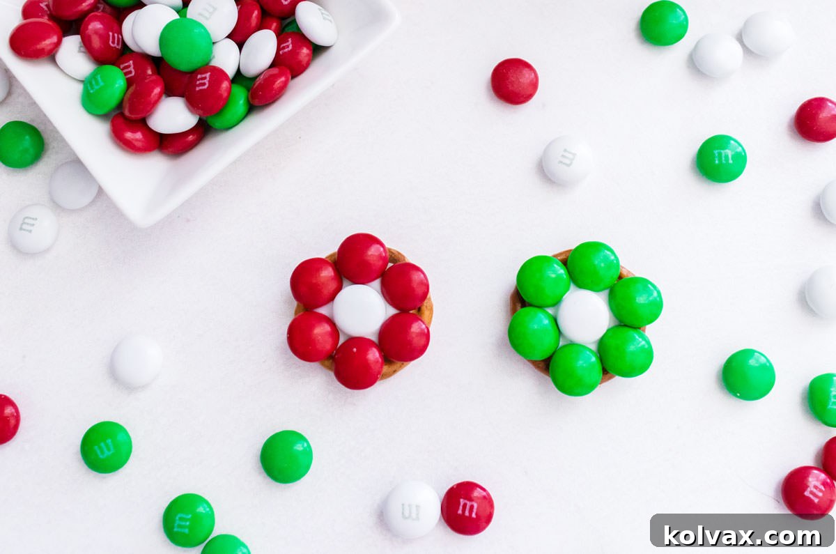 Closeup on two Christmas Flower Pretzel Bites laying on a white table surrounded by Christmas M&M's.