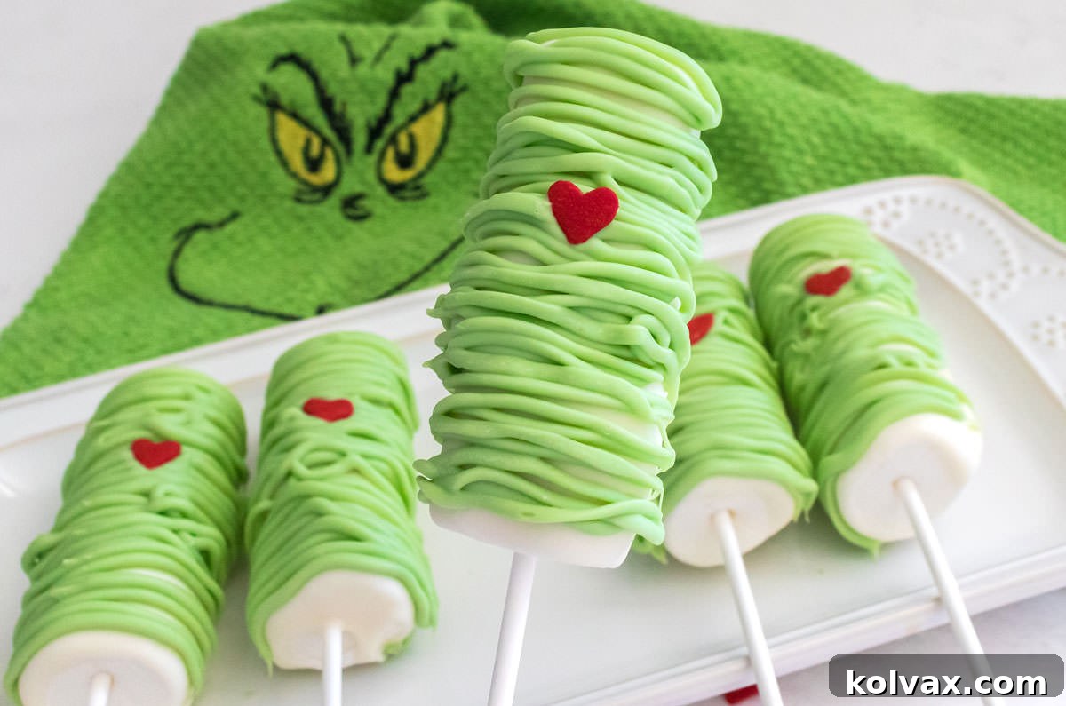 Closeup of adorable Grinch Marshmallow Pops arranged neatly on a white platter, resting on a festive Grinch-themed kitchen towel.