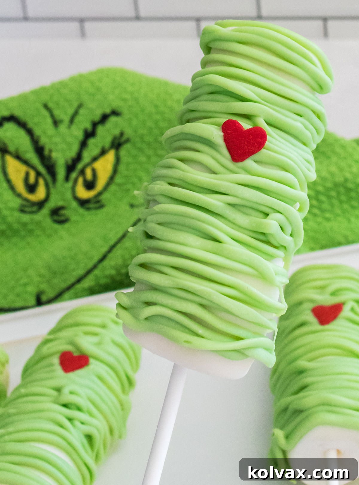 Closeup of a beautifully finished Grinch Marshmallow Pop, prominently displaying its red heart, set against a white platter and a festive Grinch kitchen towel.
