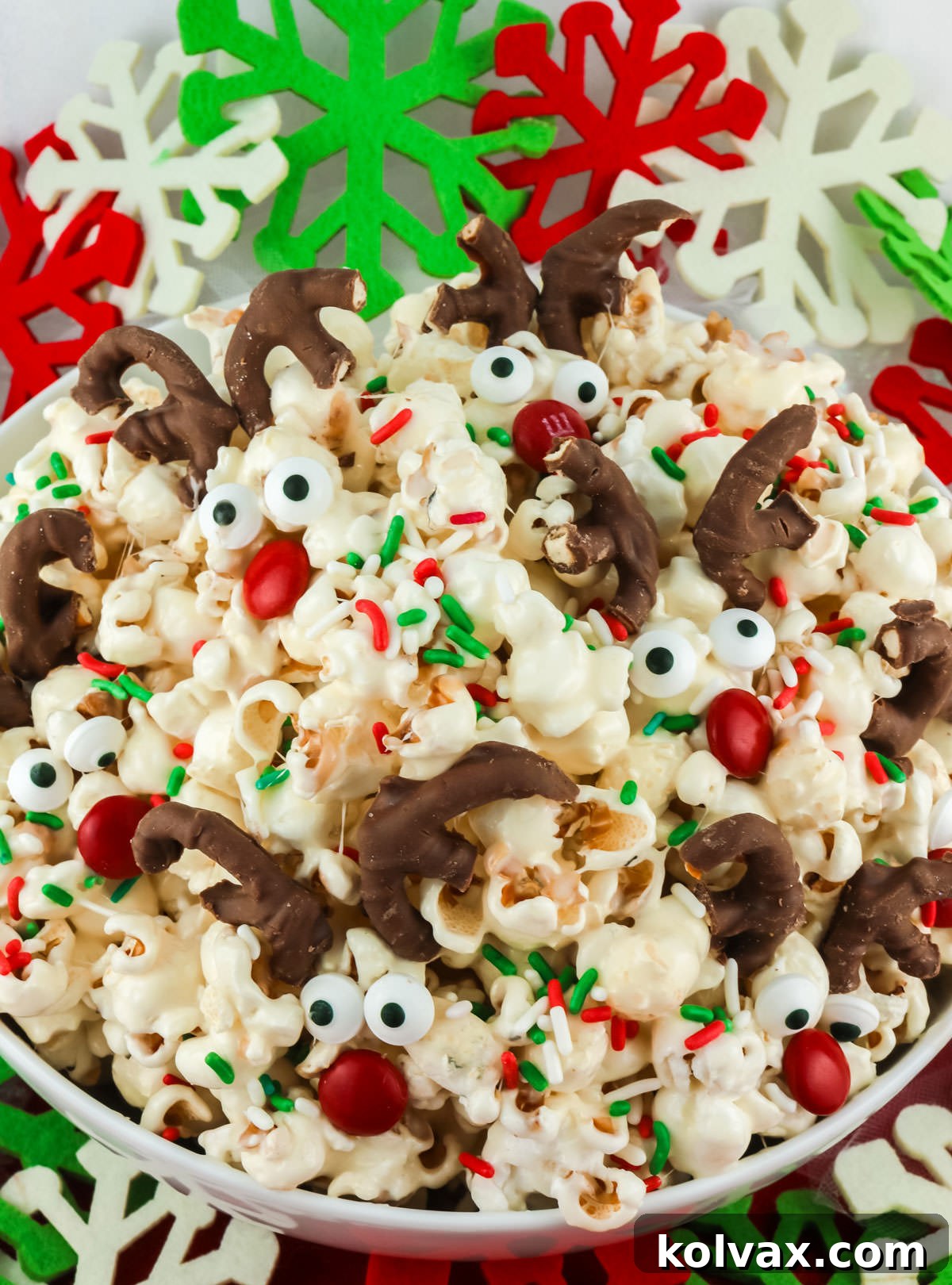 A beautiful white bowl of Reindeer Popcorn, artfully arranged with Christmas ornaments and festive greenery around it, ready for the holidays.