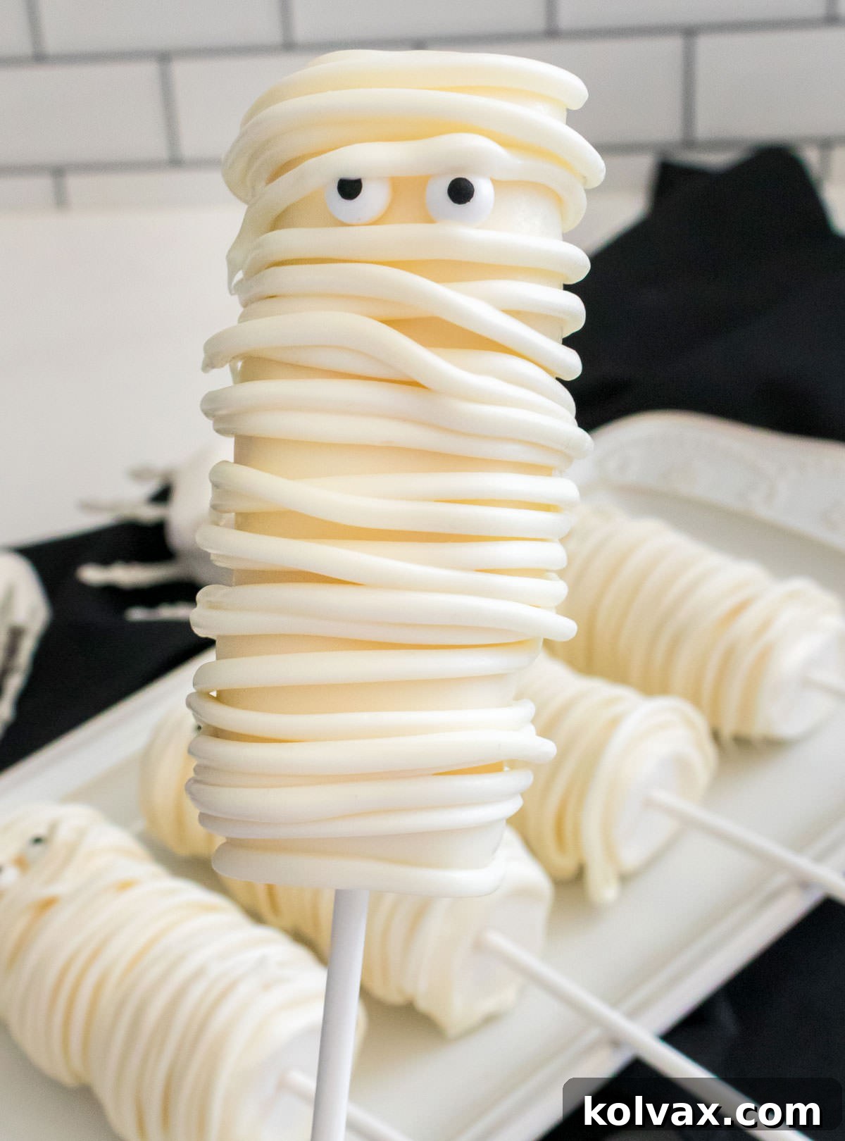 Closeup of a Mummy Marshmallow Pop hovering over a plate of more Mummy Marshmallow Pops, highlighting the finished product ready for a Halloween celebration.