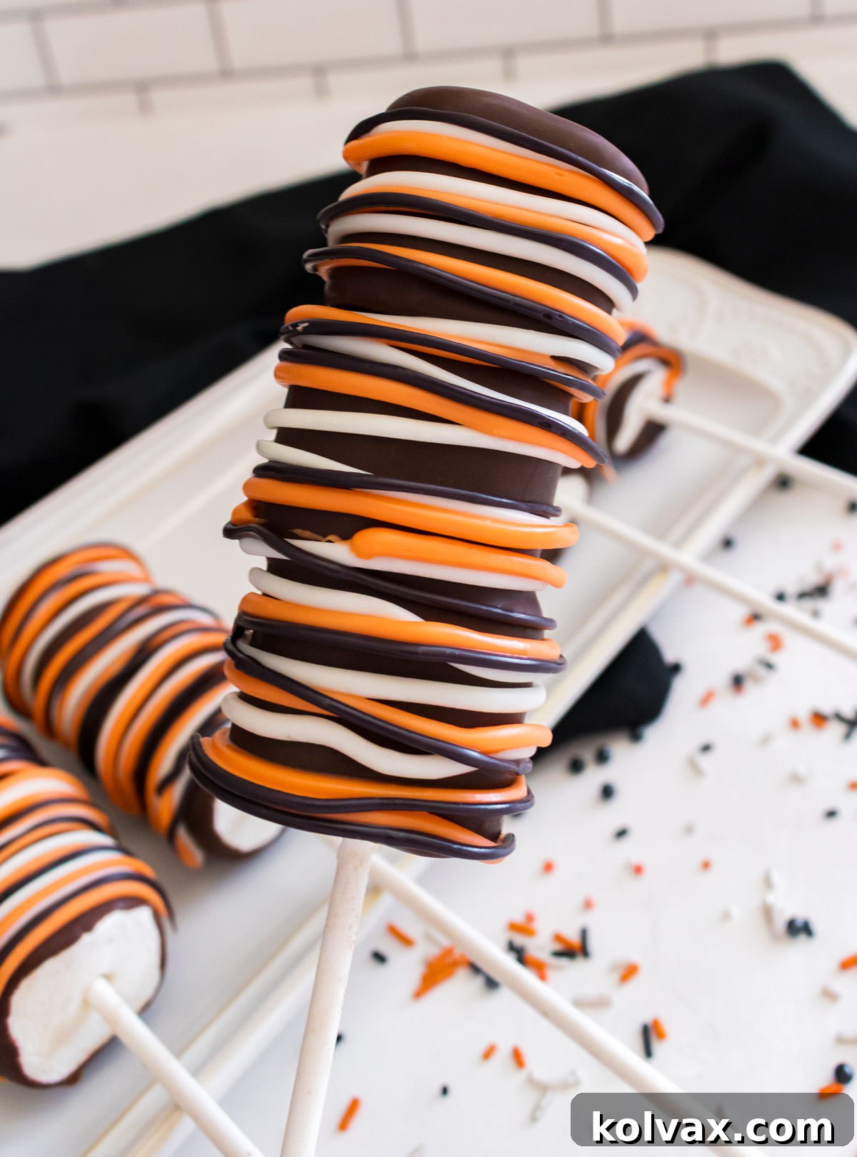 Wicked Marshmallow Wands 8 Closeup on a Halloween Marshmallow Pop hovering over a white dessert plate on a white table, showcasing the intricate drizzles and perfect finish.