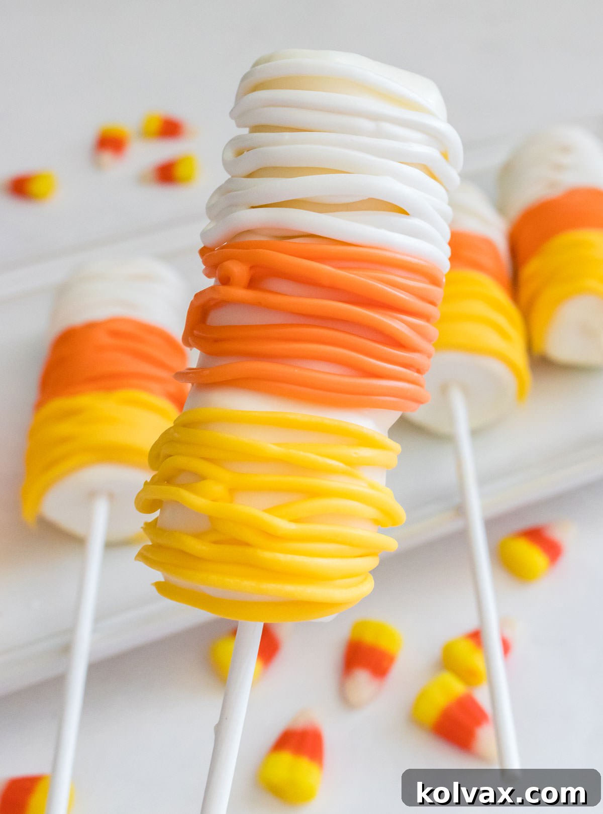 A tantalizing closeup of a perfectly crafted Candy Corn Marshmallow Pop, held gracefully above a white dessert plate. The plate is charmingly adorned with scattered candy corn candies, creating a festive backdrop on a crisp white table. This image highlights the pop's vibrant colors and appealing presentation, ideal for Halloween celebrations.