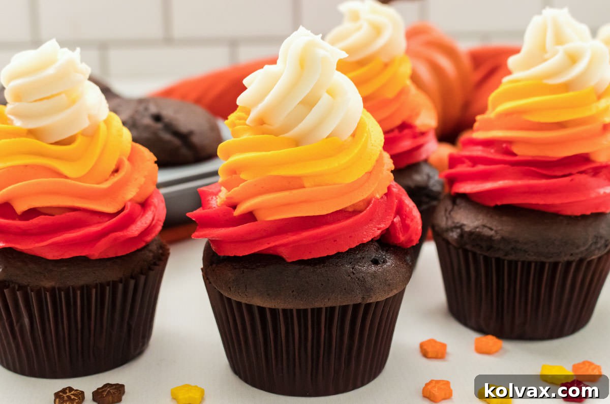 Four beautifully frosted Harvest Thanksgiving Cupcakes, featuring red, orange, gold, and white swirls, resting on a white table in front of a pan of unfrosted chocolate cupcakes.