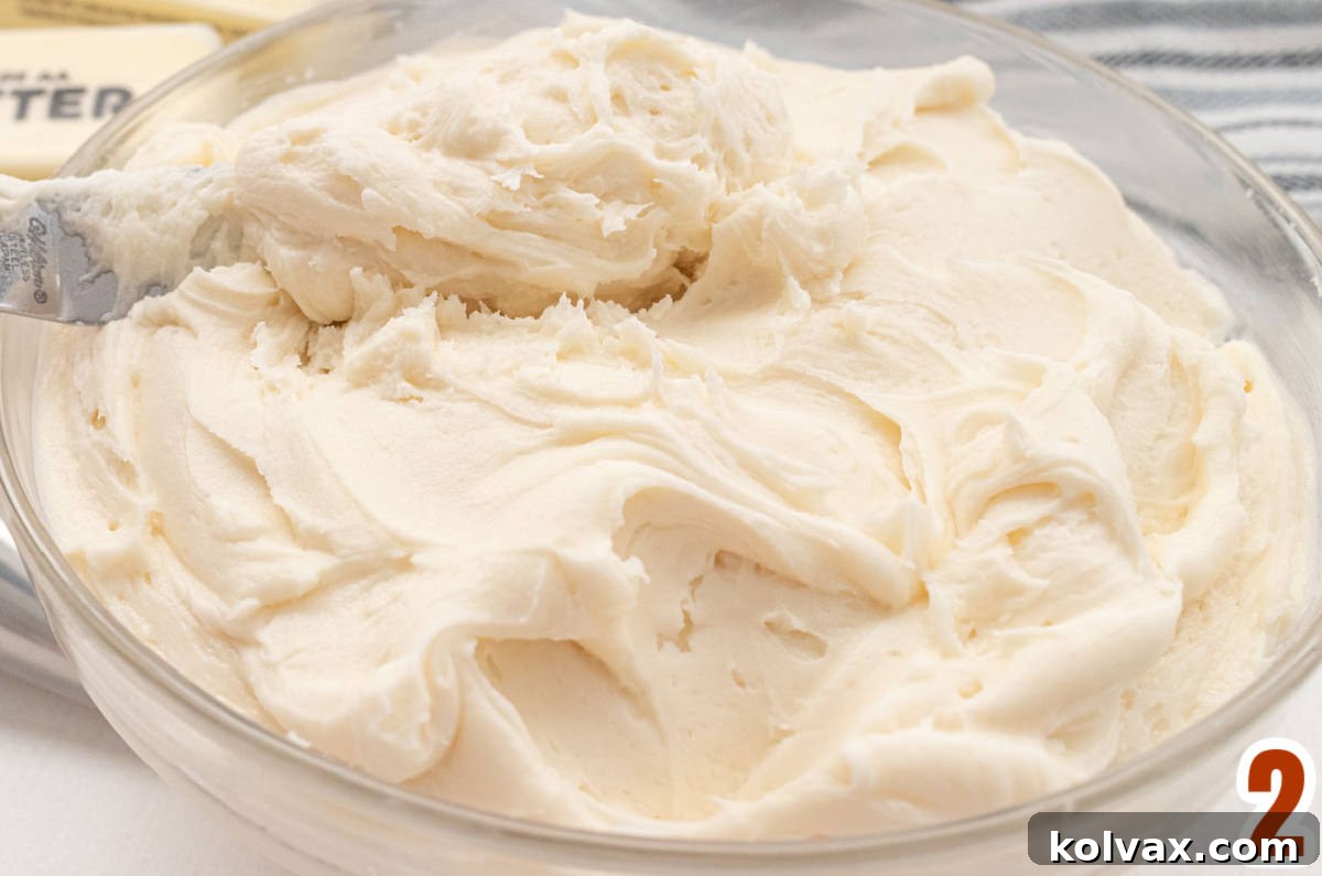 A close-up of a clear glass bowl brimming with light, fluffy, homemade Buttercream Frosting, perfectly smooth and ready for coloring.