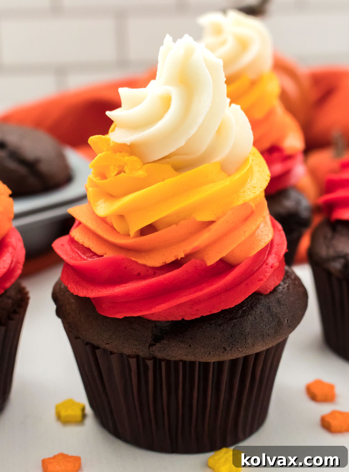 A close-up shot of a beautifully decorated Harvest Thanksgiving Cupcake, featuring a vibrant four-color swirl, resting on a white table. Other cupcakes and festive Thanksgiving sprinkles are visible in the soft-focus background.