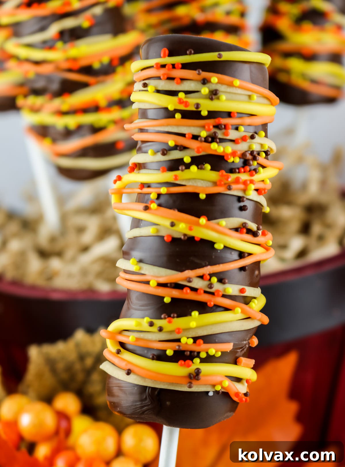 Closeup of a decorated Harvest Marshmallow Pop held against a backdrop of other vibrant candy pops, highlighting its intricate drizzle design.