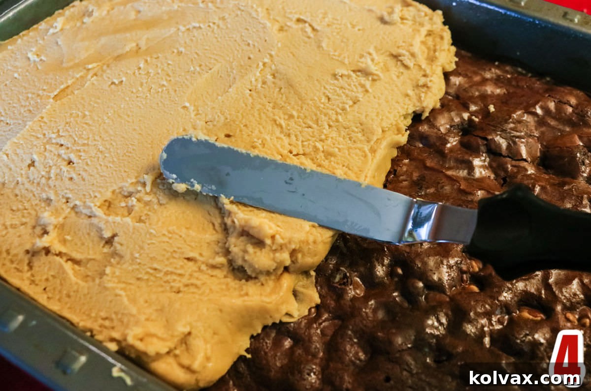 Decadent Peanut Butter Brownie Bars 6 Closeup on a pan of perfectly baked brownies being generously frosted with a thick, creamy layer of homemade Best Peanut Butter Frosting, spread smoothly across the surface with an offset spatula.