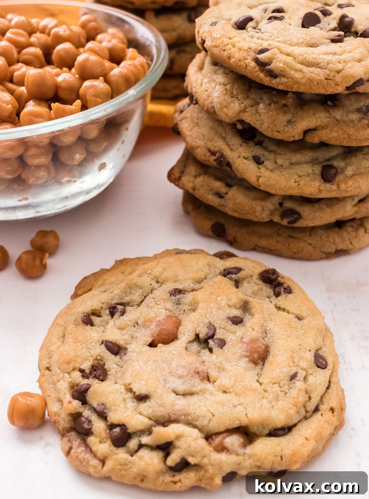 Closeup on a single Caramel Chocolate Chip Cookie sitting on a white surface in front of a stack of cookie and a glass bowl filled with Kraft Caramel Bits.