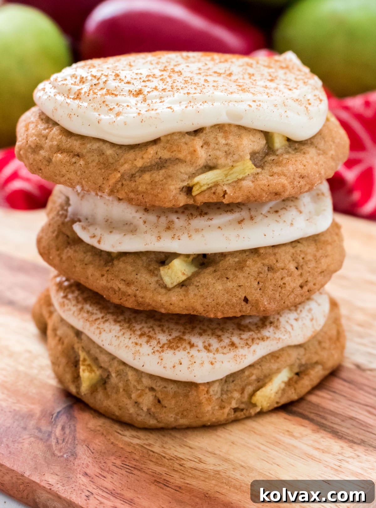 Closeup on a charming stack of three frosted Apple Cookies with Cream Cheese Frosting, artfully arranged on a rustic cutting board against a backdrop of a red towel and fresh red and green apples, emphasizing their autumnal charm.