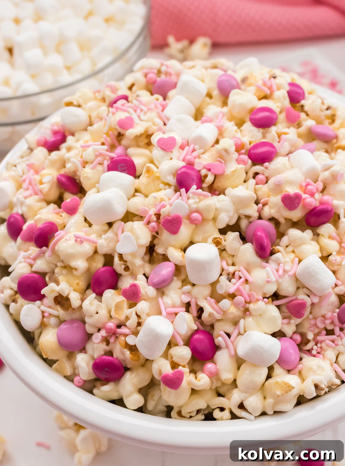 Close up of Pretty Pink Party Popcorn in a white bowl surrounded by a glass bowl filled with marshmallows and a pink table linen. 
