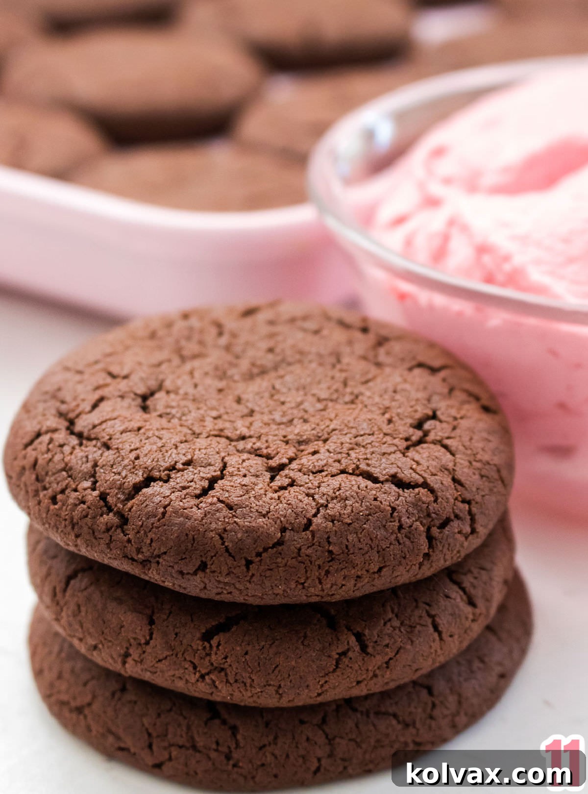 Close up of a stack of three chocolate cookies sitting next to a bowl of Raspberry Frosting and in front of a cookie sheet filled with chocolate cookies.