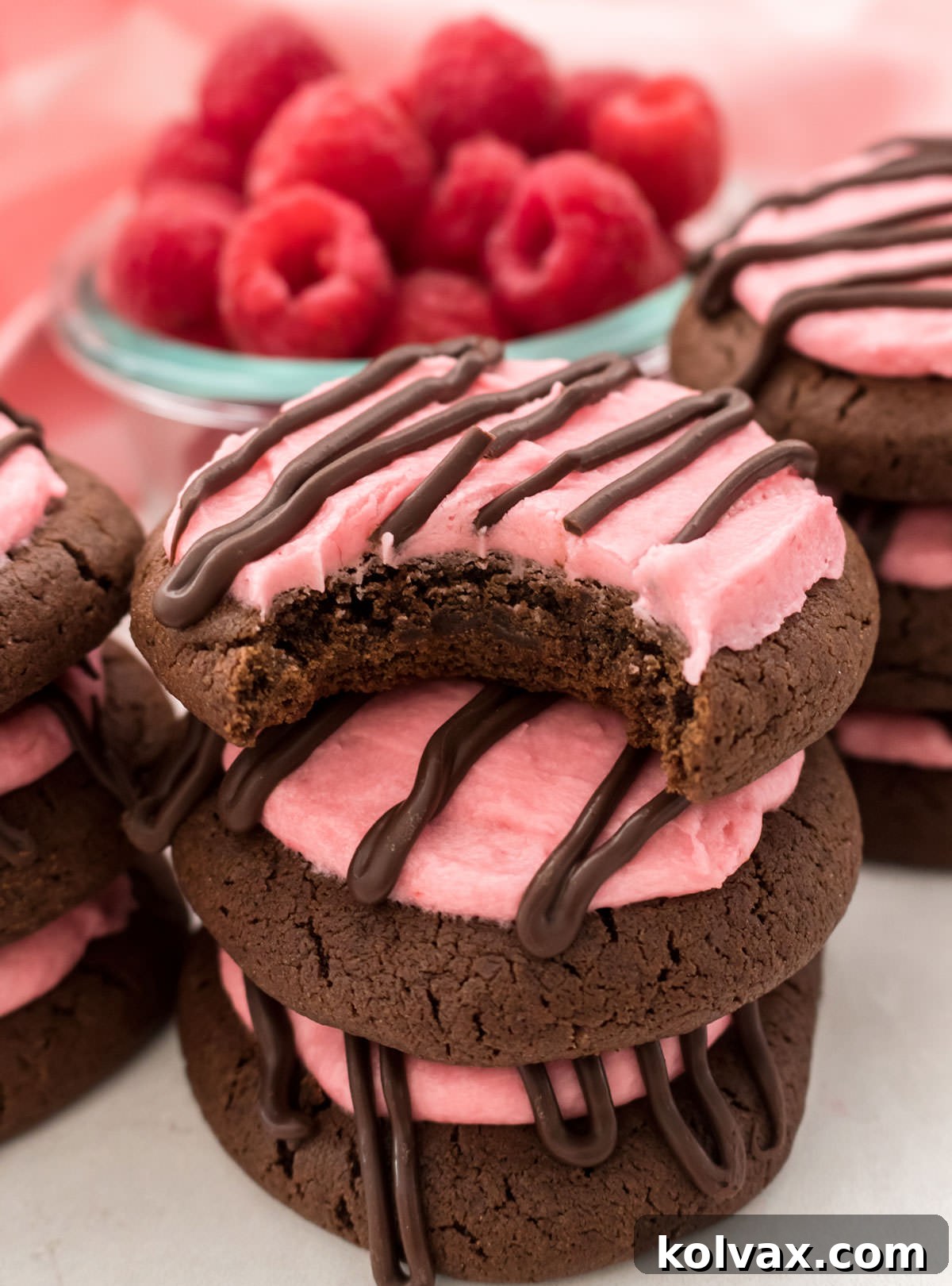 Closeup on a stack of Chocolate Cookies with Raspberry Frosting sitting on a table in front of a glass bowl filled with fresh Raspberries.