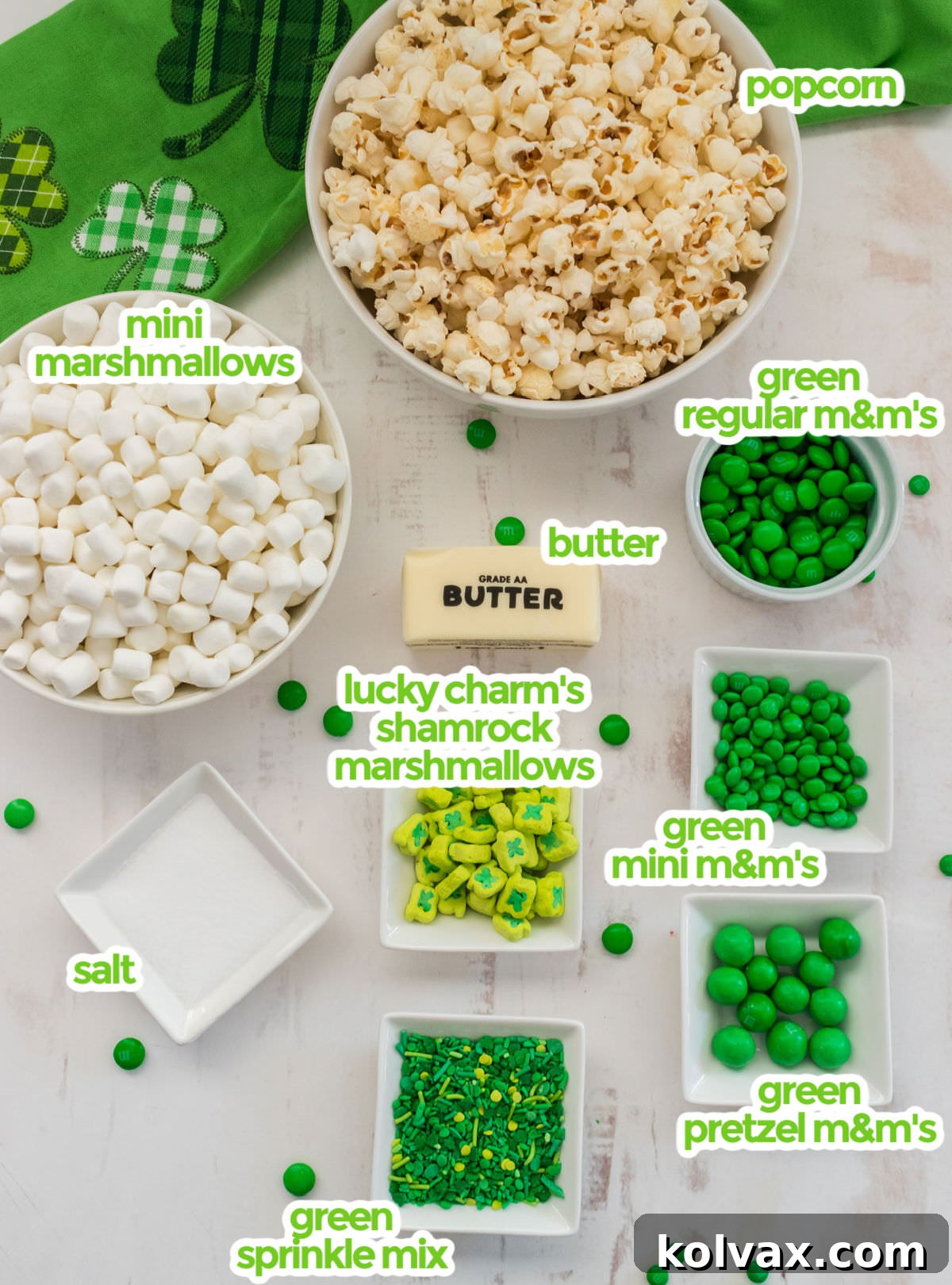 Ingredients necessary to make St. Patrick's Day Popcorn including a bowl of freshly popped popcorn, a bag of mini marshmallows, a stick of butter, a variety of green M&M's, a box of Lucky Charms for shamrock marshmallows, and a selection of green sprinkles.