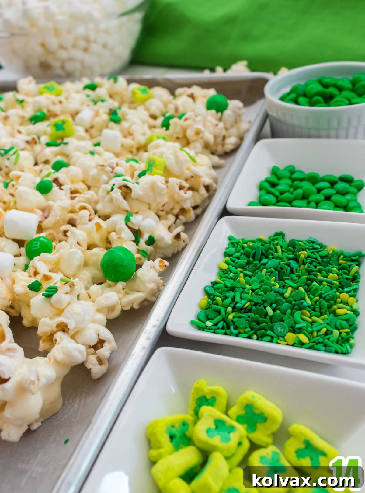 Close up on small white ramekins filled with an assortment of green St. Patrick's Day candy mix-ins, including various green M&M's, Lucky Charms shamrock marshmallows, and different green sprinkles, ready to be added to the popcorn.